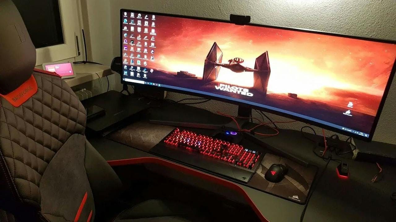 In a gaming room at a red illuminated gaming table from LeetDesk with gaming PC setup, two people stand and gamble on an Ultrawide monitor.