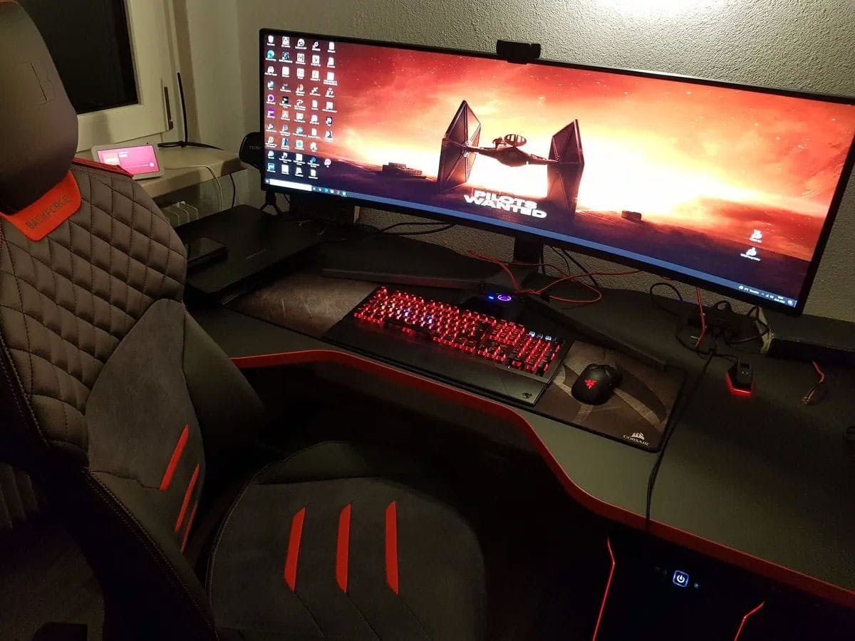 In einem Gaming Zimmer an einem rot beleuchteten Gaming Tisch von LeetDesk mit Gaming PC Setup stehen zwei Personen und zocken an einem Ultrawide Monitor