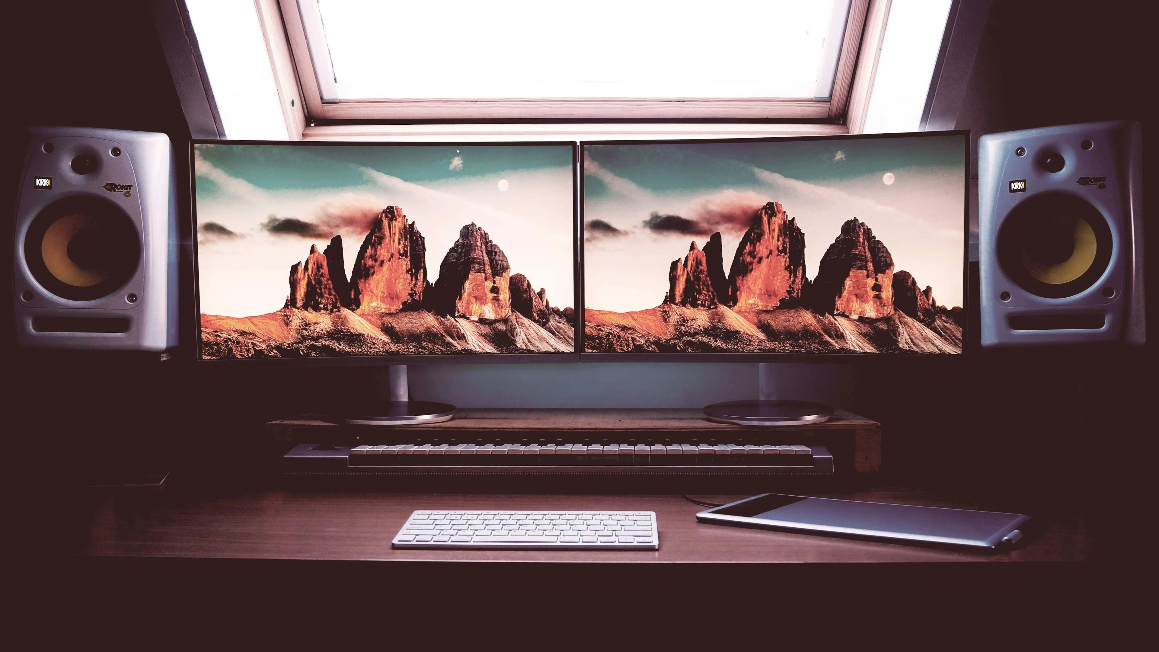 Under a window, two monitors with loudspeakers on both sides are placed frontally on a wooden table, exactly matched to each other 